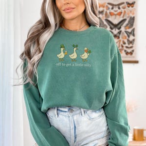 May include: A teal green sweatshirt with the phrase "off to get a little silly" printed below a graphic of three cartoon ducks. The ducks are wearing a hat, shamrocks, and holding a beer and a shamrock.
