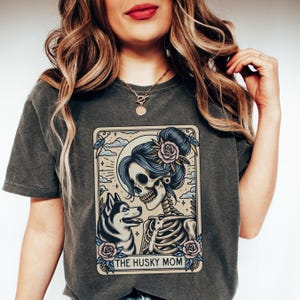 May include: Dark grey t-shirt featuring a graphic of a skeleton with blue hair and a husky dog. The design includes roses and the text "THE HUSKY MOM" in a decorative frame. The shirt is made of a soft material.