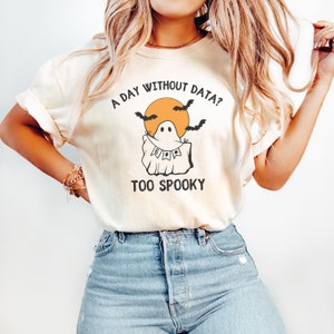 May include: Cream-colored t-shirt with a Halloween-themed graphic. The design features a ghost with a banner that says "BOO", bats, and the text "A DAY WITHOUT DATA? TOO SPOOKY".