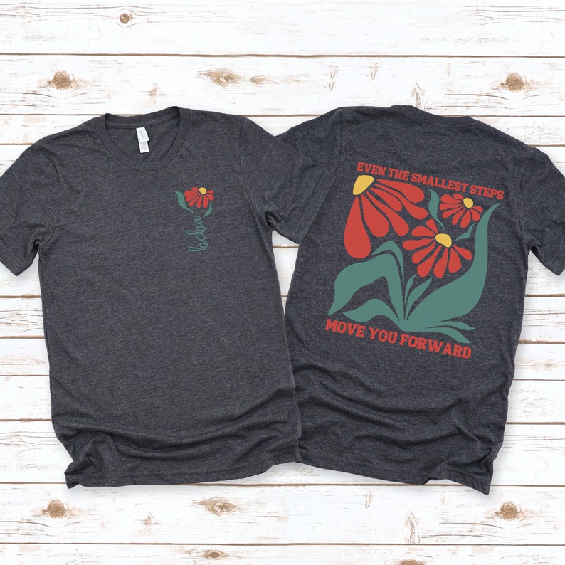 May include: A dark heather grey t-shirt with a red flower graphic on the left chest and the text "Even the smallest steps move you forward" with a red flower graphic on the back.