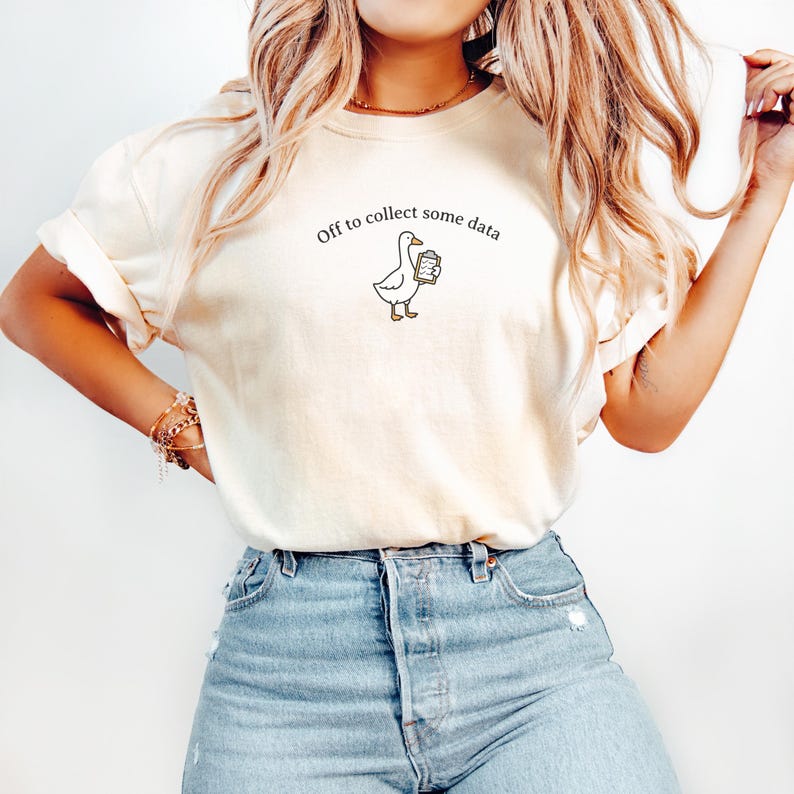 May include: Cream-colored t-shirt with the phrase "Off to collect some data" and a cartoon goose holding a clipboard. The shirt is paired with blue jeans.