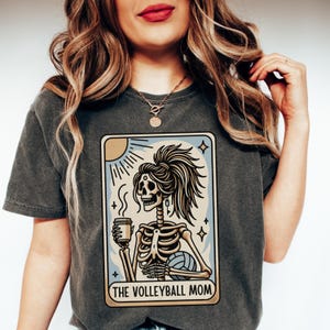 Może przedstawiać: Szary t-shirt z nadrukiem szkieletu trzymającego kubek kawy i piłkę do siatk&oacute;wki. Grafika jest w stylu karty tarota z tekstem "The Volleyball Mom" na dole.