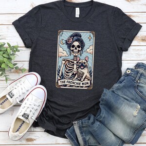May include: Dark gray t-shirt featuring a tarot card design with a skeleton, a French Bulldog, and the text "THE FRENCHIE MOM." The shirt is paired with white sneakers and blue jeans.