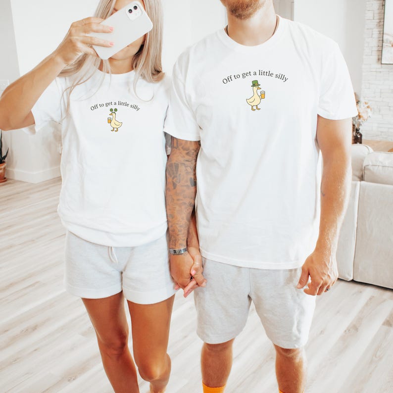 May include: White t-shirts with the text "Off to get a little silly" and a cartoon duck wearing a hat and holding a drink. The shirts are worn by two people, with one holding a phone.