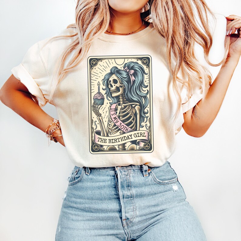 May include: A white t-shirt featuring a graphic of a skeleton holding a cupcake and a sash that reads "It's my 30th" and "The Birthday Girl".