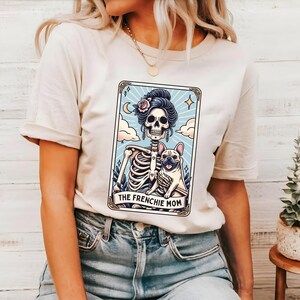 May include: Cream-colored t-shirt featuring a tarot card design with a skeleton figure holding a French Bulldog. The card has a light blue background with a crescent moon and the text "THE FRENCHIE MOM".