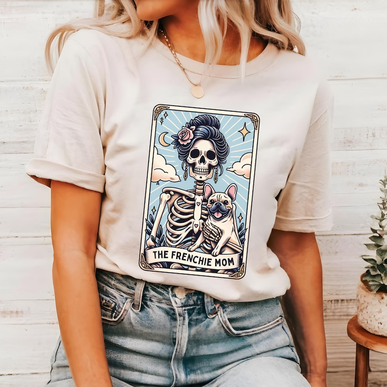 May include: Cream-colored t-shirt featuring a tarot card design with a skeleton figure holding a French Bulldog. The card has a light blue background with a crescent moon and the text "THE FRENCHIE MOM".