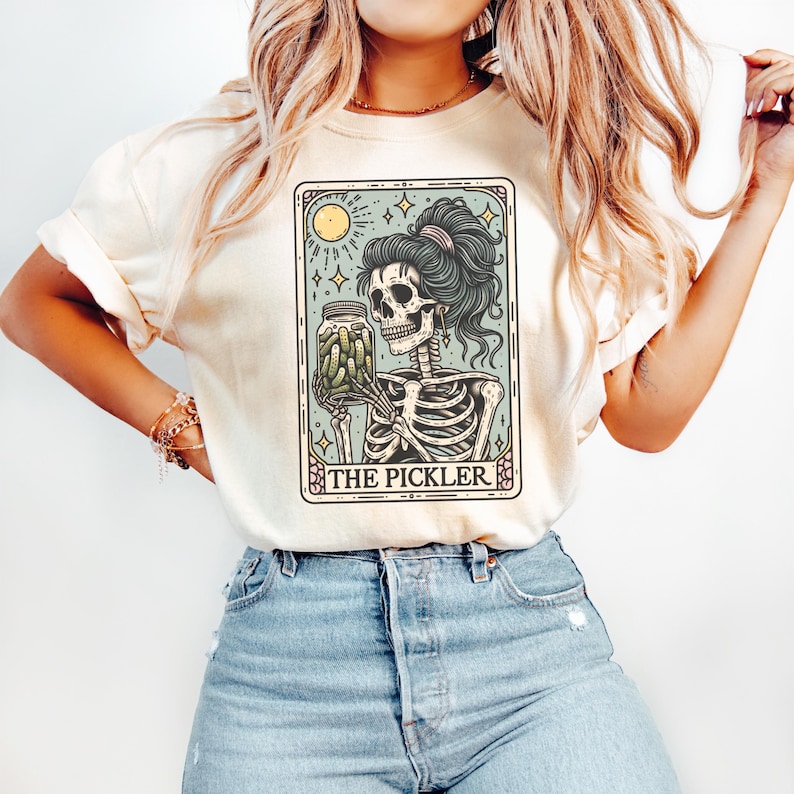Pode incluir: Uma camiseta branca com um gr&aacute;fico de um esqueleto segurando um pote de picles. O gr&aacute;fico est&aacute; no estilo de uma carta de tar&ocirc; e diz "The Pickler" na parte inferior.
