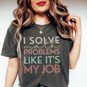 May include: Dark gray t-shirt with the text "I SOLVE math PROBLEMS LIKE IT'S MY JOB" in white, yellow, teal, and pink lettering. The shirt is a crew neck style and appears to be made of a soft material.