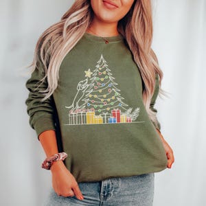 May include: An olive green sweatshirt with a white outline design of a Christmas tree, a dog, and presents. The tree is adorned with colourful lights and a star. A festive holiday-themed garment.