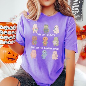 May include: A purple t-shirt with the text "THEY DID THE MATH" and "THEY DID THE MONSTER MATH". The shirt features cartoon monster characters. A person is holding an orange pumpkin mug with carved faces.