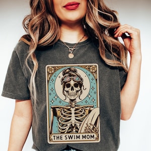May include: A dark gray t-shirt with a vintage-style illustration of a skeleton wearing sunglasses and holding a surfboard. The illustration is surrounded by a decorative frame and the text "THE SWIM MOM".