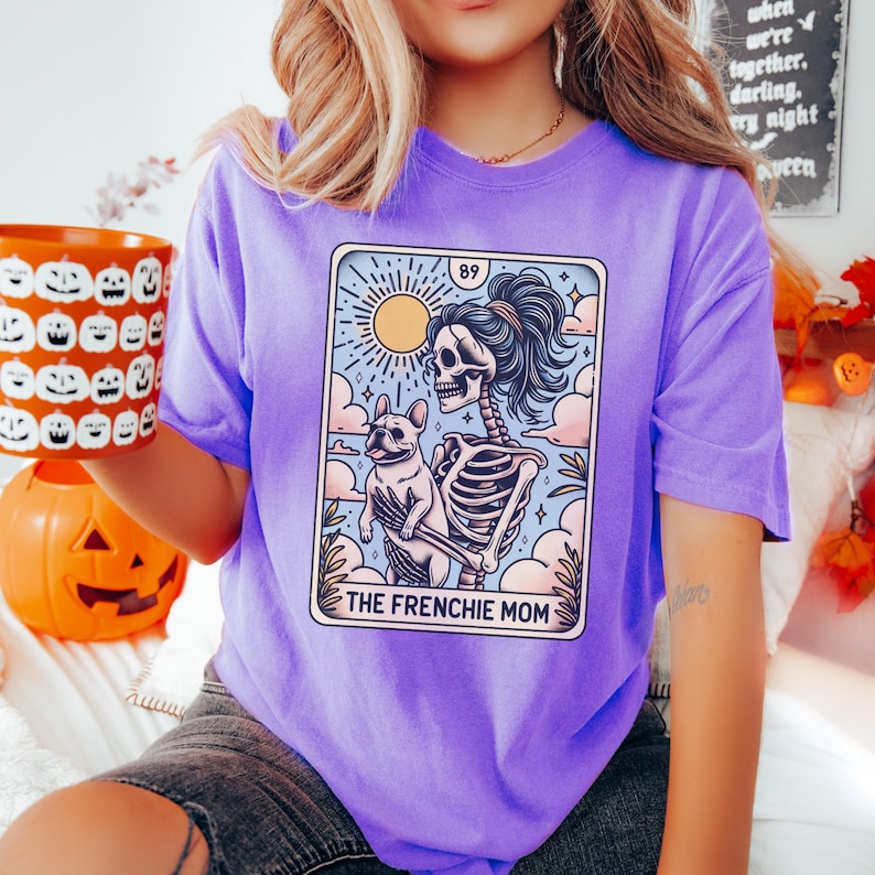 May include: Purple t-shirt with a graphic of a skeleton holding a French bulldog. The graphic is in the style of a tarot card and says "The Frenchie Mom" at the bottom.