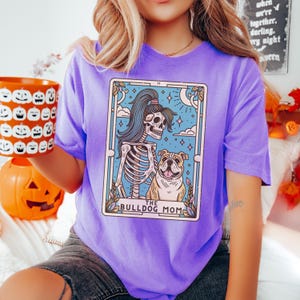 May include: Purple t-shirt featuring a graphic of a skeleton holding a bulldog. The graphic is in the style of a tarot card and reads "The Bulldog Mom".