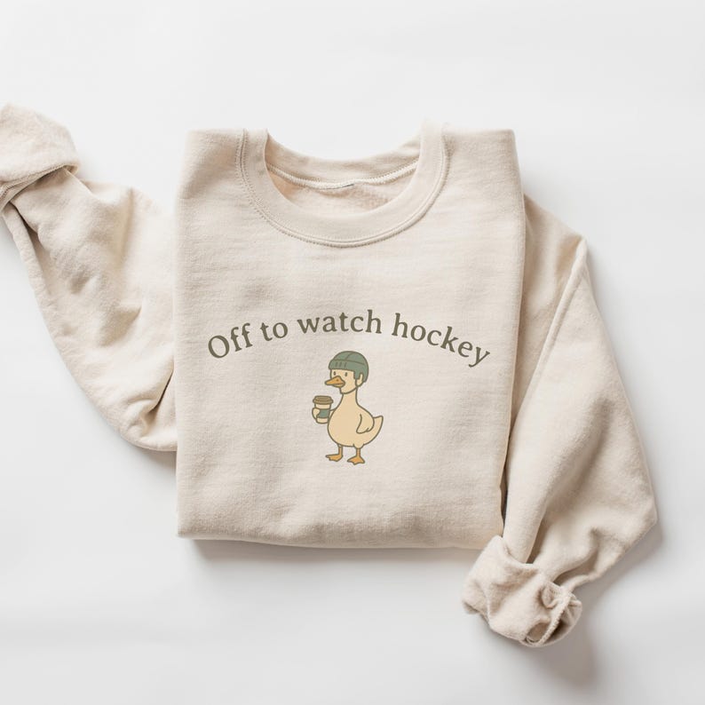 May include: A cream-colored crewneck sweatshirt featuring a graphic of a duck wearing a green hockey helmet and holding a coffee cup. The text "Off to watch hockey" is printed in dark gray lettering above the duck illustration on the chest.