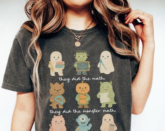 May include: Dark grey t-shirt featuring nine cartoon monsters, each holding a math-related item. The text "they did the math" and "they did the monster math" are printed below the monsters. The shirt is a comfortable, casual style.