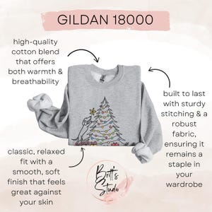 May include: A light grey Gildan 18000 sweatshirt with a Christmas tree design. The sweatshirt has a classic, relaxed fit and is made of a high-quality cotton blend. The design includes a dog decorating a Christmas tree with colourful lights.