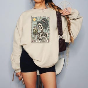 May include: Beige sweatshirt featuring a tarot card-style graphic. The graphic depicts a skeleton holding a jar of pickles, with the text "THE PICKLER" at the bottom. The design includes a sun, moon, and stars.