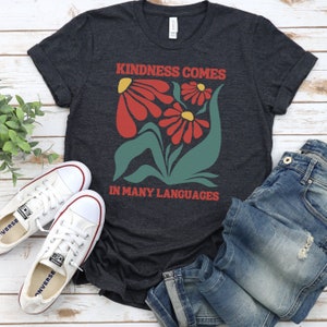 May include: A dark grey t-shirt with a graphic design of red flowers and green leaves. The text "Kindness comes in many languages" is printed on the shirt.