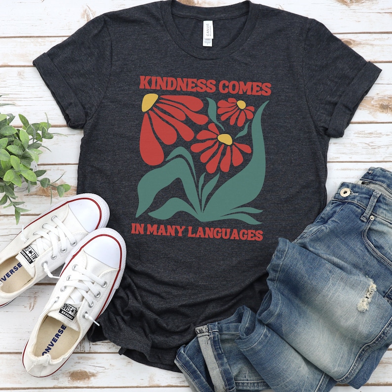 May include: A dark grey t-shirt with a graphic design of red flowers and green leaves. The text "Kindness comes in many languages" is printed on the shirt.