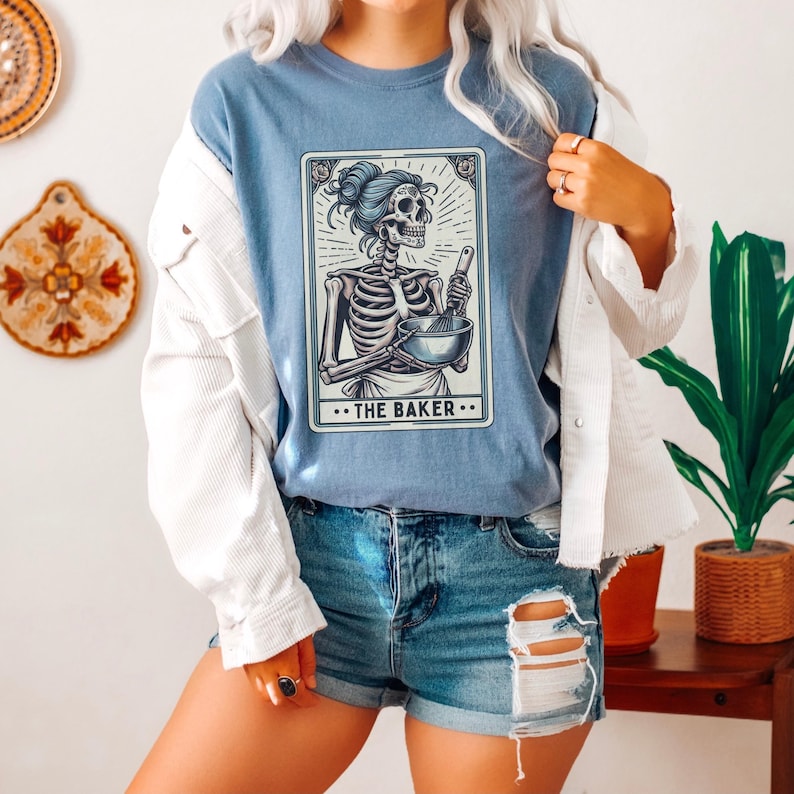 May include: A blue t-shirt with a graphic of a skeleton holding a mixing bowl and a whisk. The graphic is in a tarot card style with the text "...THE BAKER..." at the bottom.