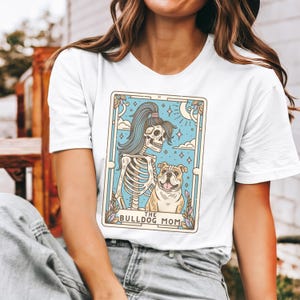 May include: White t-shirt featuring a graphic of a skeleton with a ponytail holding a bulldog. The graphic is in a tarot card style with the text "The Bulldog Mom".