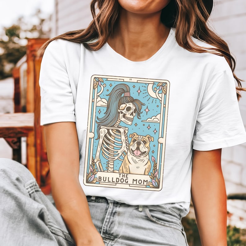 May include: White t-shirt featuring a graphic of a skeleton with a ponytail holding a bulldog. The graphic is in a tarot card style with the text "The Bulldog Mom".
