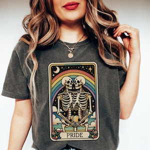 May include: A gray t-shirt with a tarot card design featuring two skeletons holding hands in front of a rainbow. The card reads "PRIDE" at the bottom.