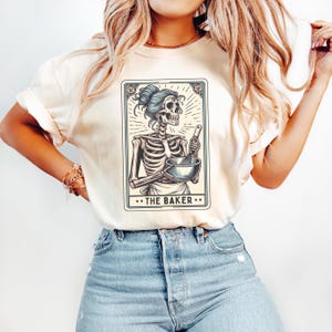 May include: A white t-shirt with a graphic of a skeleton holding a bowl and whisk. The graphic is in a tarot card style with the text "...THE BAKER..." at the bottom.