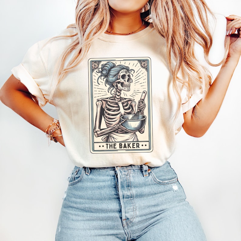 May include: A white t-shirt with a graphic of a skeleton holding a bowl and whisk. The graphic is in a tarot card style with the text "...THE BAKER..." at the bottom.