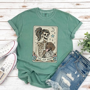 May include: A green t-shirt with a vintage-style illustration of a skeleton holding a dog. The illustration is in a card-like frame with the text "The GSP Mom".