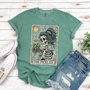 Pode incluir: Uma camiseta verde com um design de carta de tar&ocirc; vintage que apresenta um esqueleto segurando um frasco de picles. O cart&atilde;o diz "The Pickler".
