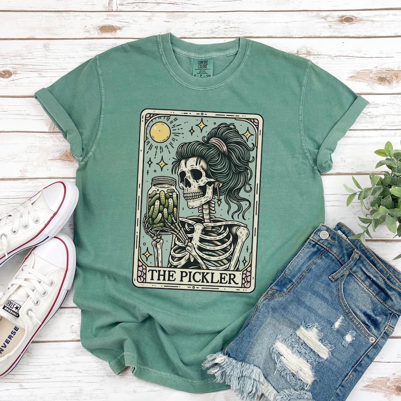 Pode incluir: Uma camiseta verde com um design de carta de tar&ocirc; vintage que apresenta um esqueleto segurando um frasco de picles. O cart&atilde;o diz "The Pickler".