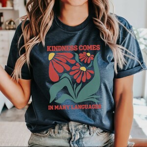 May include: A navy blue t-shirt with a graphic of red flowers and green leaves. The text "Kindness Comes In Many Languages" is printed on the shirt.