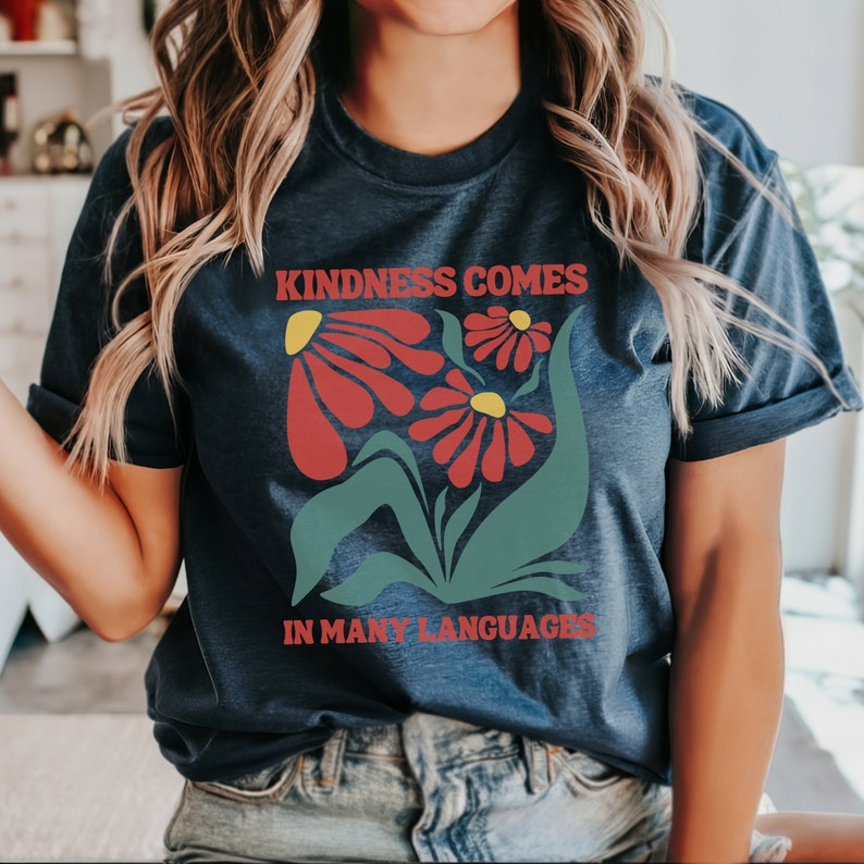 May include: A navy blue t-shirt with a graphic of red flowers and green leaves. The text "Kindness Comes In Many Languages" is printed on the shirt.