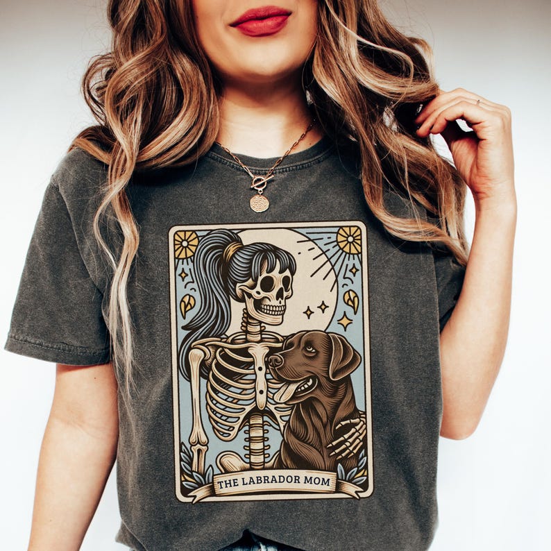 May include: A dark gray t-shirt with a vintage-style illustration of a skeleton holding a labrador retriever. The illustration is surrounded by a border with the text "THE LABRADOR MOM".