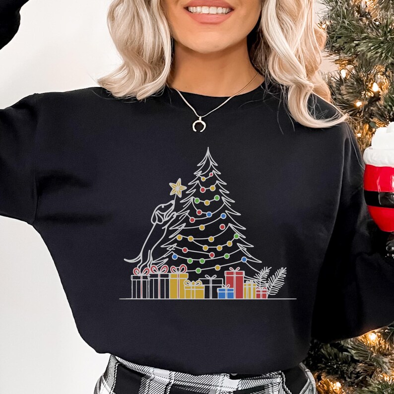 May include: Black sweatshirt with a white outline design of a Christmas tree, a dog, and gifts. The tree has colourful lights and a star. A festive design for the holiday season.