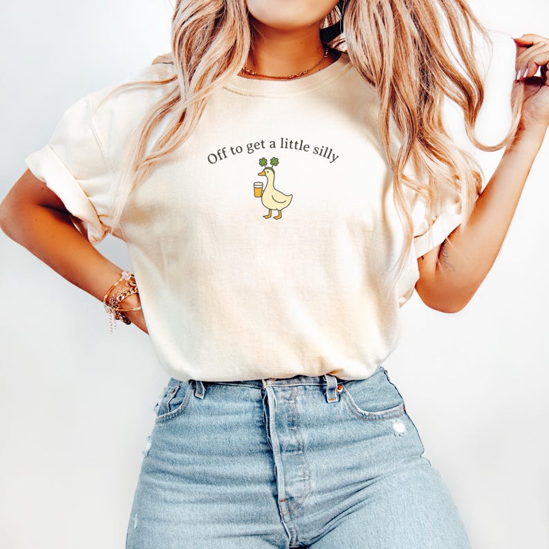 May include: Cream-colored t-shirt with the text "Off to get a little silly." The shirt features a cartoon duck holding a beer and wearing a shamrock. The shirt is paired with blue jeans.