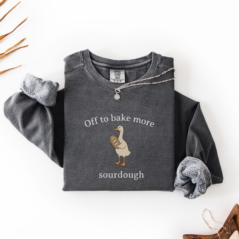 Puede incluir: Sudadera de manga larga gris oscuro con el texto "Off to bake more sourdough" y un dibujo de un ganso sosteniendo pan. Un collar plateado con un colgante peque&ntilde;o est&aacute; sobre la sudadera. Las mangas est&aacute;n dobladas.