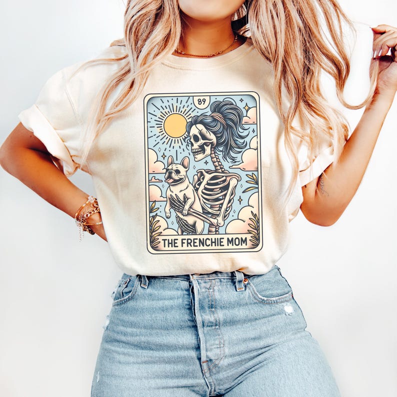 May include: A white t-shirt with a graphic of a skeleton holding a French bulldog. The graphic is in the style of a tarot card and says "The Frenchie Mom" at the bottom.