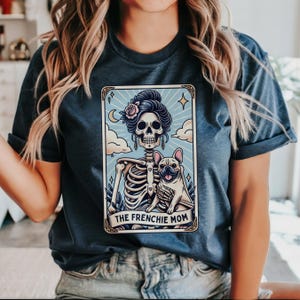 May include: Dark blue t-shirt featuring a tarot card design with a skeleton, a French Bulldog, and the text "THE FRENCHIE MOM". The card has a light blue background with clouds, a crescent moon, and stars.