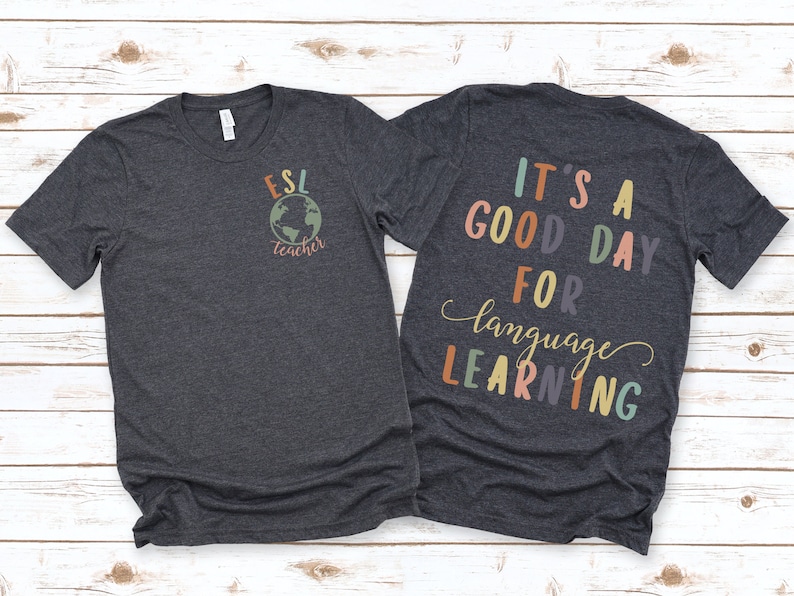 May include: A dark heather grey t-shirt with the text "ESL teacher" and a globe graphic on the front. The back of the shirt has the text "It's a good day for language learning" in colorful letters.