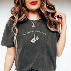 May include: Dark gray t-shirt with the phrase "Off to collect some data" above a cartoon goose holding a clipboard. The goose is white with yellow feet and a yellow beak. The shirt is a casual style.