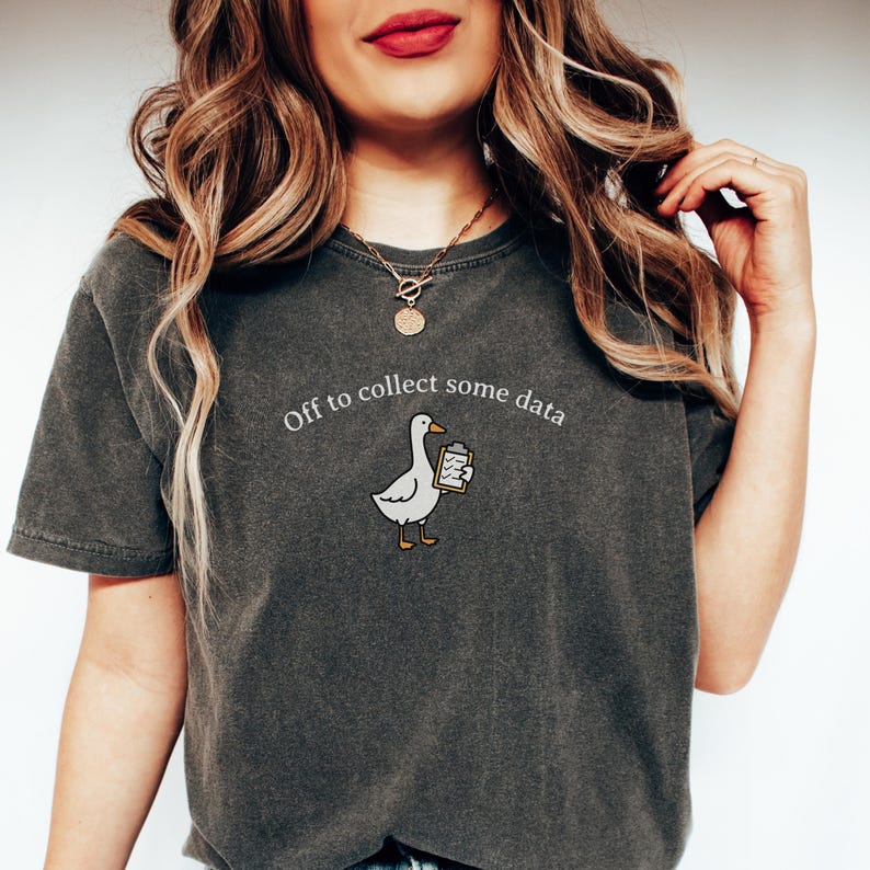 May include: Dark gray t-shirt with the phrase "Off to collect some data" above a cartoon goose holding a clipboard. The goose is white with yellow feet and a yellow beak. The shirt is a casual style.