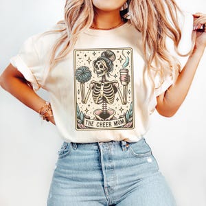 Pode incluir: Uma camiseta branca com um gr&aacute;fico de um esqueleto segurando uma x&iacute;cara de caf&eacute; e um pompom de torcida. O gr&aacute;fico &eacute; de estilo vintage e tem o texto "The Cheer Mom" na parte inferior.