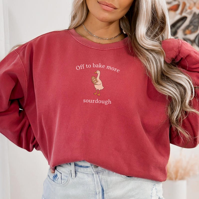 Puede incluir: Una sudadera granate con el texto "Off to bake more sourdough" y un gr&aacute;fico de un ganso sosteniendo un pan. La sudadera es de color liso y tiene cuello redondo.