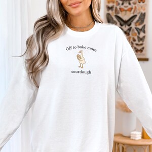 Puede incluir: Sudadera blanca con el texto "Off to bake more sourdough" y un dibujo de un ganso con pan. La sudadera es de manga larga y cuello redondo.