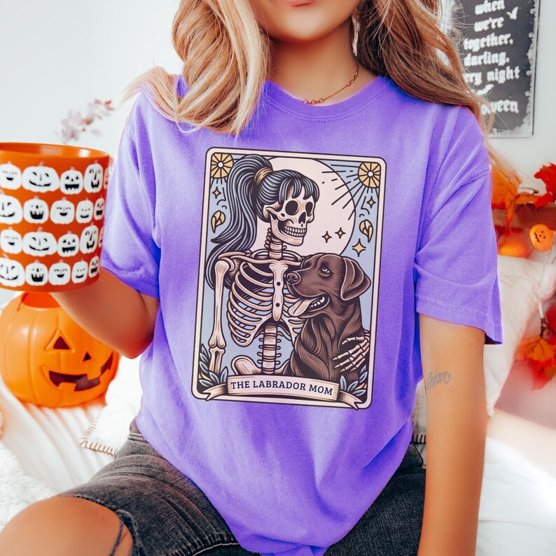 May include: Purple t-shirt with a graphic of a skeleton holding a labrador retriever. The graphic is in a tarot card style with the text "The Labrador Mom" at the bottom.
