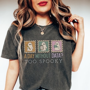 May include: Dark gray t-shirt featuring three postage stamp designs with ghost illustrations. The stamps are in yellow, teal, and purple. Text on the shirt reads "A DAY WITHOUT DATA? TOO SPOOKY."