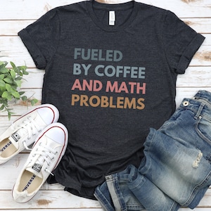 May include: A dark heather grey t-shirt with the text "Fueled by Coffee and Math Problems" in colorful block letters. The shirt is laid out on a light wood surface with a pair of white canvas shoes and blue jeans.